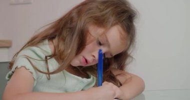 Liseli kız, egzersiz kitabında kalemle yazma işi yapıyor. Uzun saçlı küçük kız cam masada oturur ve ev ödevlerini yakın plan yapar.