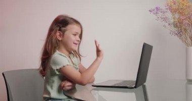 Küçük kız bilgisayardan arkadaşınla konuşurken el sallıyor. Sevimli çocuk, vazonun yanındaki çiçekli cam masada otururken hoş bir jest gösteriyor.