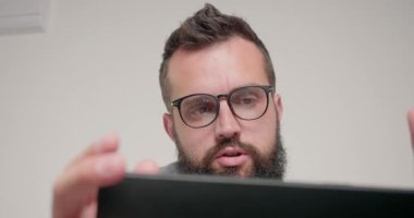 Gözlüklü sakallı adam bilgisayarla müdürle görüntülü görüşme yapıyor. Serbest Çalışan, odada bej duvara karşı oturarak yeni görevi tartışıyor