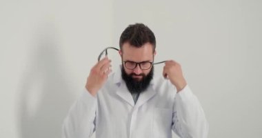 Gözlüklü doktor gülümser ve kameraya bakarak steteskop takar. Beyaz önlüklü sakallı terapist klinikte her zamanki çalışma gününe başlamaya hazır.