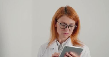 Gözlüklü kızıl saçlı kadın doktor tablet üzerinde çalışıyor. Doktor hastalara çevrimiçi danışmanlık yapıyor. Ciddi bayanın muayenehanede her zamanki çalışma günü.