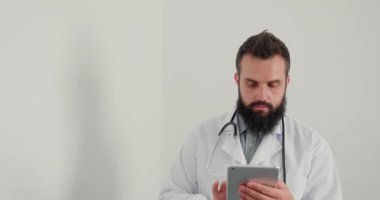 Erkek doktor klinikte tablet üzerinde çalışıyor. Sakallı terapist, internet üzerinden hastalara e-reçeteli danışmanlık yapıyor. Ciddi adam kameraya bakıyor.