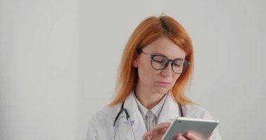 Kızıl saçlı kadın doktor tablet üzerinde çalışıyor. Tıp çalışanı hastalara çevrimiçi danışmanlık yapıyor. Odaklanmış kadın klinikteki uzaklığa bakar ve düşünür.