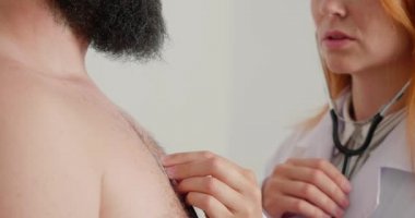Kadın doktor klinikteki hastayla çalışıyor. Genel pratisyen, yetişkin bir adamın ciğerlerini steteskopla inceliyor. Kızıl saçlı bayan konsültasyonda.