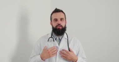 Steteskoplu doktor gülümseyip kameraya bakarak tavsiye veriyor. Doktor hastalara online tavsiye veriyor. Beyaz önlüklü sakallı terapistin klinikte her zamanki işi var.. 