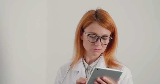眼鏡の赤い髪の女性の医師は タブレット上で動作します 医者は患者にオンラインで相談する 深刻な女性は 通常の勤務日で医師事務所 — ストック動画