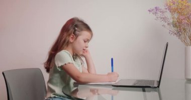Küçük kız, egzersiz kitabında görev yaparken dizüstü bilgisayarla online eğitim alıyor. Çalışkan ortaokul öğrencisi cam masaya oturur ve kalemle yazar.