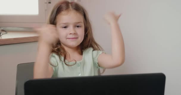 快乐的女孩在完成家庭作业后表现出大拇指向上的姿态 心满意足的小女孩坐在写字台旁 挨着开着的笔记本电脑 在家里的特写镜头前 — 图库视频影像