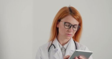 Kızıl saçlı kadın doktor tablet üzerinde çalışıyor. Tıp çalışanı hastalara çevrimiçi danışmanlık yapıyor. Odaklanmış kadın klinikteki uzaklığa bakar ve düşünür.