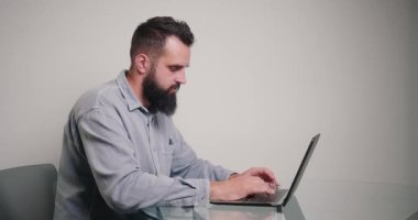 Genç metin yazarı, dizüstü bilgisayardaki makaleyi kameraya çeviriyor. Klasik tişörtlü, sakallı esmer adam masa başında oturmuş, ofisten internet üzerinden çalışıyor.