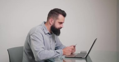 Adam online ödeme yapıyor ve kredi kartının detaylarını dizüstü bilgisayarla giriyor. Genç sakallı çalışan işyerinde internetten ürün alıyor.