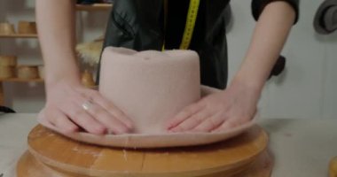 Experienced milliner straightens felt hat brim on wooden mold at table in workshop. Woman works with elastic material creating headwear closeup