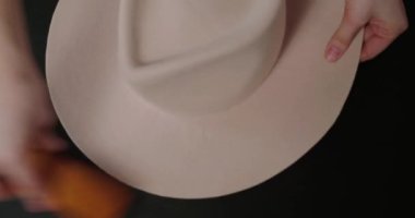 Milliner brushes felt hat standing at black table on grey background. Woman takes care of handmade female headwear in workshop closeup