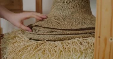 Woman takes wicker straw hats standing on wooden shelf. Professional milliner creates stylish female headwear for sale in workshop closeup