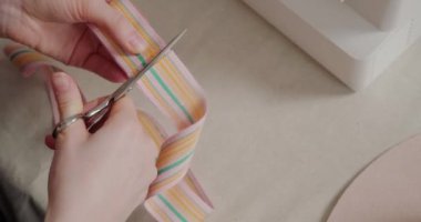 Woman cuts wide colorful ribbon with scissors sitting at white table. Skillful milliner prepares decor for felt hat in workshop closeup