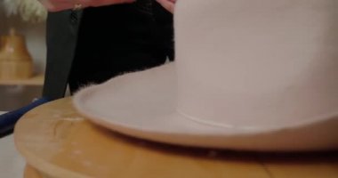 Woman measures brim of felt hat on wooden mold using yellow tape-line. Professional milliner makes stylish headwear in workshop closeup
