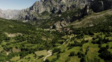 Bare rocky mountains and hills covered with trees and grass surround canyon with small village on sunny day. Long road runs in highland aerial view