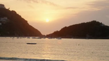 Tourist yachts sail in sea against giant forestry mountains at bright sunset. Sun reflects on sea water surface with waves rolling on beach