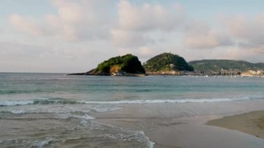 Light sea waves with blue water roll on shore washing soft yellow sand. Empty beach in coastal town against forestry mountains at sunset