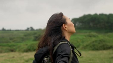 Young Asian woman enjoys windy weather standing with closed eyes on blurred background. Hiker with backpack in wild area closeup slow motion