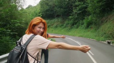 Young women show hitchhiking hand gesture to stop passing car turning to camera and smiling. Hikers walk on road in wilderness closeup slow motion