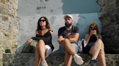 Friends sit on ancient building stone parapet talking and using gadgets. Man and women in sunglasses rest after hiking on sunny day slow motion