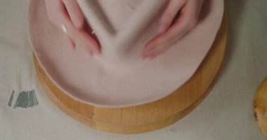 Hands of woman stretch material of felt hat to make brim shape. Milliner finishes working with headwear on round wooden mold in workshop closeup