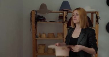 Tatmin olmuş şapkacı kameraya bakarak şapkasını takıyor. Genç sarışın kadın kendi atölyesinde elinde şık el yapımı başlıkla duruyor.