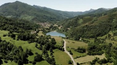 Road runs on hills with green grass past blue lake in highland valley on sunny summer day. Giant forestry mountains covered with lush trees aerial view