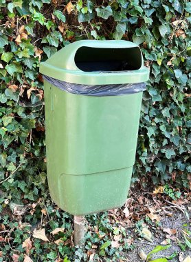 Vertical shot on a green plastic public wastebasket for disposal of garbage against a wall with green ivy