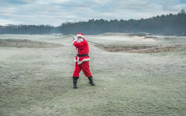 Kırmızı geleneksel kostümlü Noel Baba, kışın dışarıda, yeşil bir buz tarlasında golf oynar..