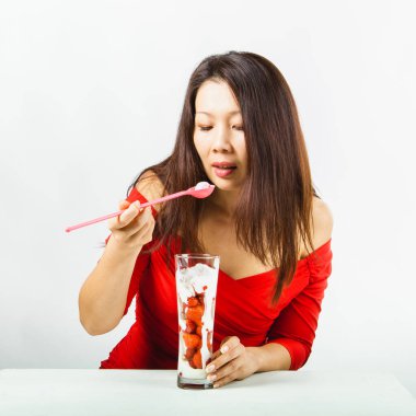 Kırmızı elbise giyen Asyalı güzel kadın lezzetli çilek meyveli tatlı yiyor. Stüdyo çekimi, beyaz arka plan, ön manzara.