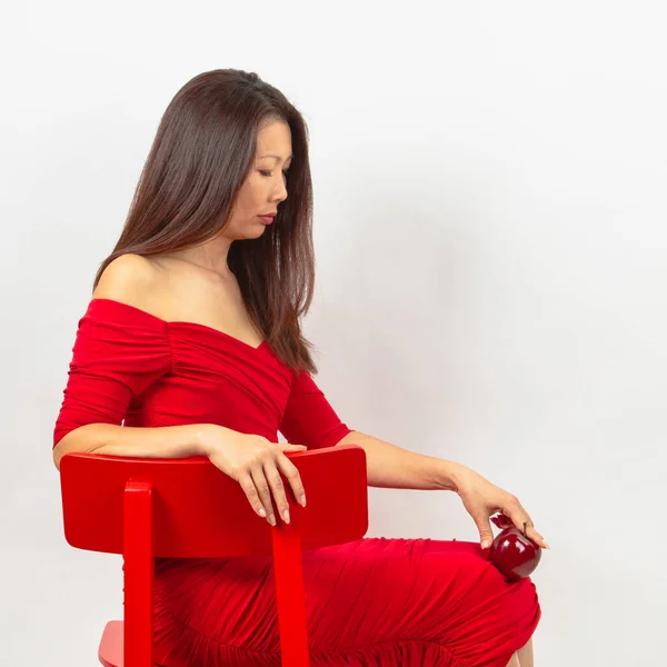 エレガントなアジアの女性 長い髪のブルネットは ネックラインの赤いドレスで赤い椅子に座って 彼女の手に赤いリンゴを持っています 白い背景で撮影されたスタジオ — ストック写真