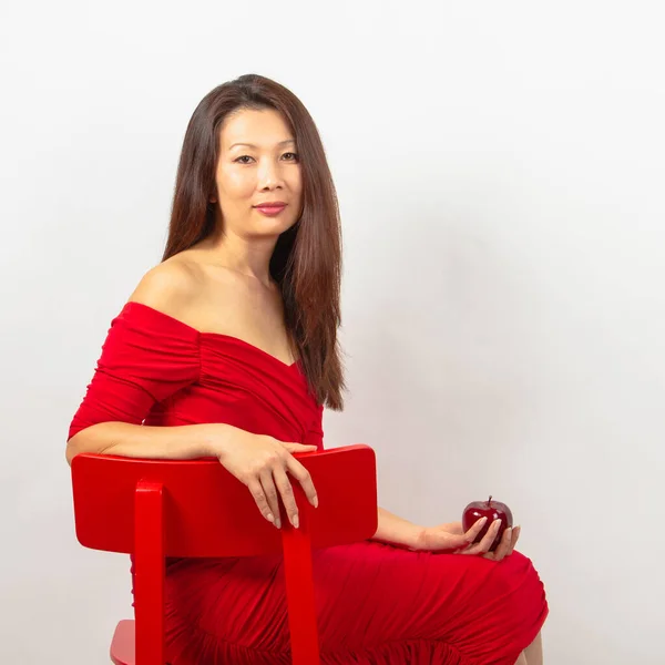 美しいアジアの女性 長い髪のブルネットは ネックラインの赤いドレスで赤い椅子に座って 彼女の手に赤いリンゴを持っています 白い背景で撮影されたスタジオ — ストック写真