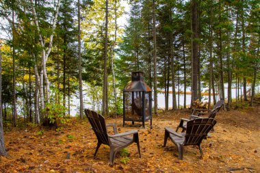 Gölün kenarındaki ormanda şezlongların ve şöminenin olduğu güzel bir sonbahar sahnesi.