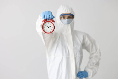 Koruyucu tıbbi giysili ve maskeli kadın beyaz arka planda kırmızı alarm saati tutuyor.