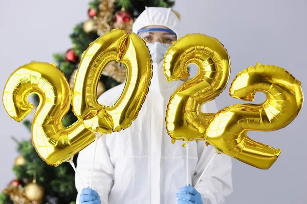 Женщина в защитном костюме и маске держит номера 2022 на фоне рождественской елки — стоковое фото