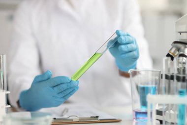 Koruyucu eldivenler giyen bilim adamı yeşil sıvıyla test tüpü tutuyor.
