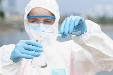 Koruyucu kıyafetli ve gözlüklü bilim adamı deney tüpünden şişeye su döküyor.