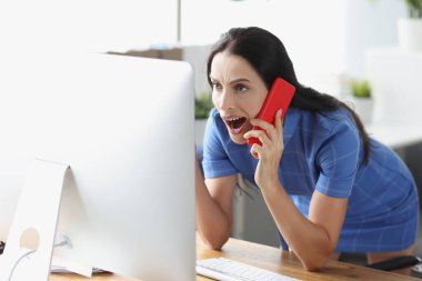 Şok olmuş genç bir kadın akıllı telefondan konuşuyor ve bilgisayar ekranına bakıyor.