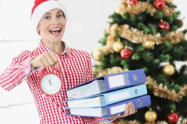 Портрет молодой улыбающейся женщины в шляпе Санта-Клауса держит будильник и деловые документы на заднем плане — стоковое фото