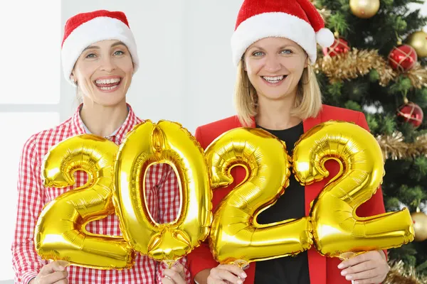 Две смеющиеся девушки в шляпах Санта-Клауса, держащие новогодние номера 2022 — стоковое фото