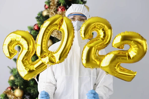 Врач в защитном костюме, маске и очках держит номера 2022 на фоне новогодней елки — стоковое фото