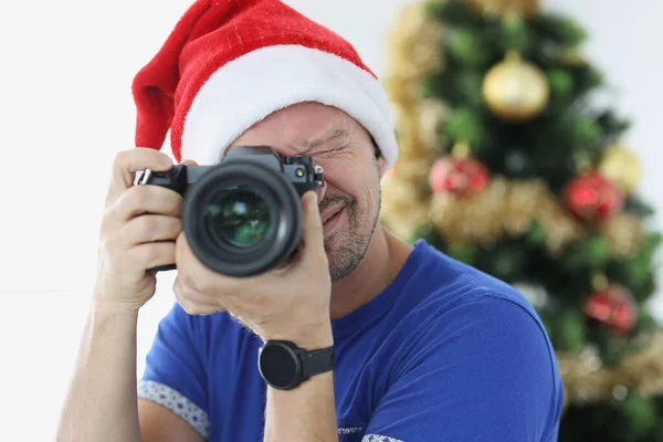 Mężczyzna w czerwonym kapeluszu Mikołaja robi zdjęcia aparatem w pobliżu choinki — Zdjęcie stockowe