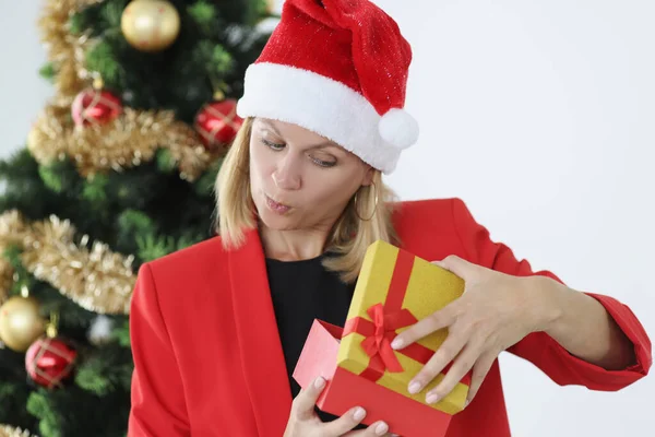 Mujer joven en rojo Santa Claus sombrero apertura regalo de año nuevo cerca del árbol y se sorprende —  Fotos de Stock