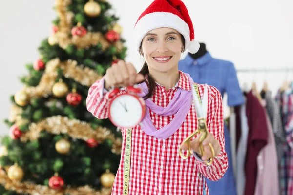 Seamstress in red Santa hat holding alarm clock and scissors near Christmas tree — Stock Photo, Image