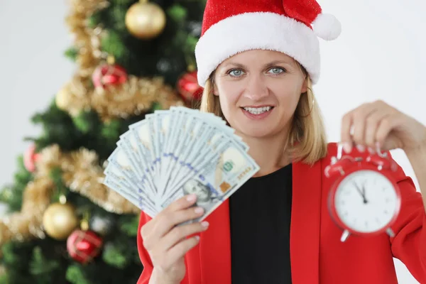 Kırmızı Noel Baba şapkalı kadın yeni yıl ağacının yanında elinde para ve çalar saat tutuyor. — Stok fotoğraf