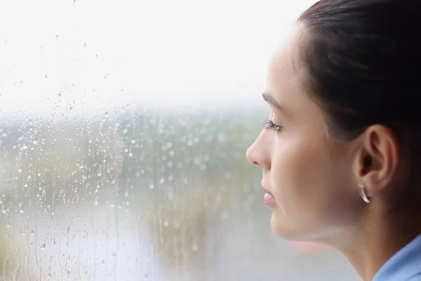 Νεαρή σκεπτική γυναίκα κοιτάζει έξω από το παράθυρο στη βροχή — Φωτογραφία Αρχείου