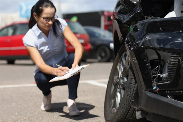 Inspektorin schreibt Dokumente auf Klemmbrett neben kaputtem Auto. Autoversicherung — Stockfoto