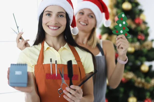 Две женщины-парикмахерши в шляпах Санта-Клауса держат ножницы и расчески с календарем на фоне новогодней елки — стоковое фото